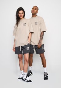 T-shirt oversize beige con logo sul petto, abbinata a pantaloni corti neri con cordoncino e logo. Entrambi indossano sneakers e calze.