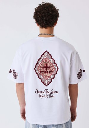 Persona con capelli ricci che indossa una maglietta bianca con un motivo decorativo bordeaux e la scritta "Change The Game Project X Paris" sul retro.