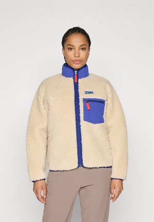Kvinne iført beige fleecejakke med blå krage og brystlomme, stående mot en enkel lys bakgrunn.