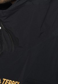 Veste en tissu noir avec un col haut, dotée d'une fermeture éclair et d'une petite poche. Le logo est imprimé en doré. Texture lisse, design minimal.