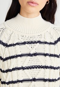 Kremfarget høyhalset genser med teksturert kabelstrikk og marineblå striper. Stoffet har et mykt, chunky design med hevede mønstre.