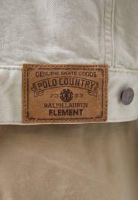 Læderetiket syet på beige stof, der står "Genuine Skate Goods Polo Country 2023 Ralph Lauren Element" med en metalknap i nærheden.