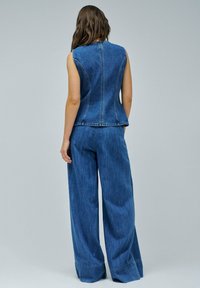 Gilet en denim avec un design ajusté sans manches, associé à un pantalon en denim à jambes larges. Les deux sont dans un lavage bleu moyen avec des coutures visibles.