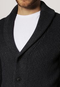 Mörkgrå ribbad cardigan med en sjalkrage, prydd med två svarta knappar, bärs över en vit t-shirt. Minimalistisk design och textur.