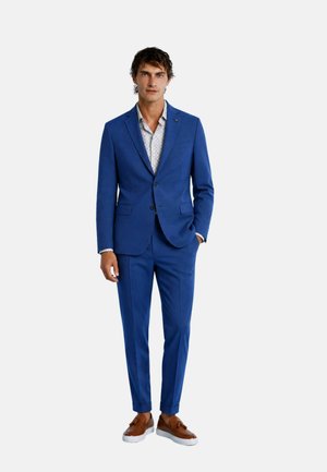 Uomo in piedi che indossa un completo blu con camicia bianca a fantasia e mocassini marroni con nappine su sfondo bianco uniforme.
