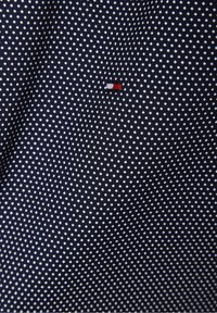 Tessuto blu navy con piccoli pois bianchi e un piccolo logo rettangolare ricamato rosso e bianco vicino al centro.