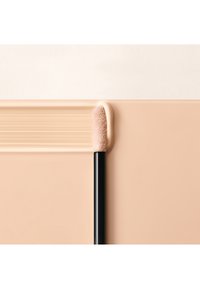 Aplicador de labios con una punta suave y esponjosa, mango negro y una muestra de producto beige claro y suave sobre un fondo neutro.
