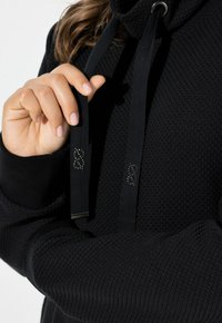 Osoba trzymająca sznurki od czarnej bluzy z kapturem z symbolami ampersand, ubrana w teksturowany czarny sweter z prążkowanymi mankietami.