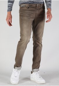 Vaqueros ajustados de color marrón con una textura desgastada, con un bolsillo frontal, y combinados con zapatillas blancas, con los puños doblados hacia arriba para revelar un forro gris.