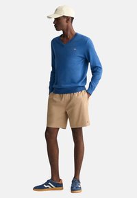 Blauer V-Ausschnitt-Pullover, beige Shorts und blaue Sneakers mit orangefarbenen Akzenten; trägt eine hellfarbige Mütze. Glatte Textur und lässige Passform.
