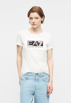 Νεαρό άτομο με κοντά ανοιχτό καστανά μαλλιά φοράει λευκό t-shirt EA7 Emporio Armani και ανοιχτόχρωμο τζιν, στέκεται μπροστά σε απλό φόντο.