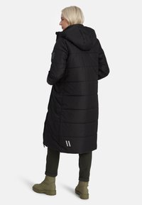 Melnas polsterētās jakas ar kapuci, vertikāli izšūtas dizains, atstarojoši akcenti un pagarināts siluets, saskaņots ar zaļiem biksēm un zābakiem.