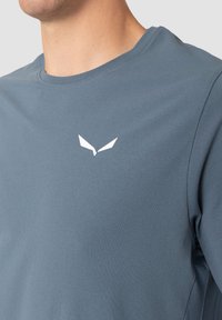 Grå T-skjorte med korte ermer laget av mykt stoff, med en hvit logo på brystet. Rund hals, glatt tekstur og enkelt design.