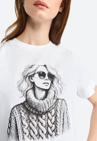 T-shirt bianco con un'illustrazione in bianco e nero di una donna che indossa occhiali da sole e un maglione a collo alto con motivo. Maniche corte, collo rotondo.