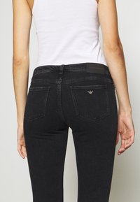 Musta farkkukangas housut kapealla leikkauksella, joissa on takataskut ja merkkipainatus nahkapätkä vyötäröllä. Kangas on sileä.