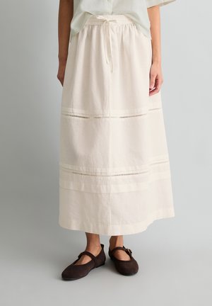 Jupe blanche longueur cheville avec taille à cordon et bandes horizontales décoratives, portée avec des chaussures Mary Jane marron foncé sur fond gris.