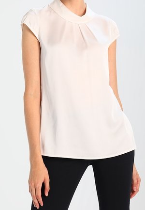 Femme portant un chemisier blanc sans manches avec un col rond et un pantalon noir sur un fond uni.