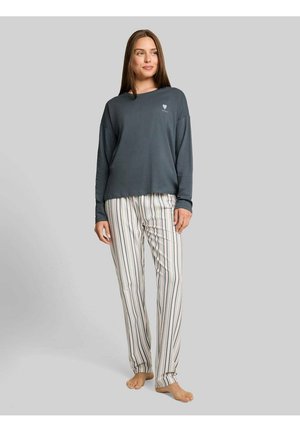 Vrouw staat op blote voeten, draagt een donker top met lange mouwen en witte broek met verticale zwarte en beige strepen op een grijze achtergrond.