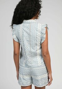 Haut en tissu bleu clair avec des broderies blanches complexes, des manches courtes à volants et des motifs géométriques, associé à un short assorti.