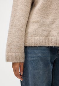 Béžový, textúrovaný pletený sveter s rebrovanými manžetami, kombinovaný s modrými džínami. Zamerajte sa na mäkký materiál a jemné farebné odtiene.