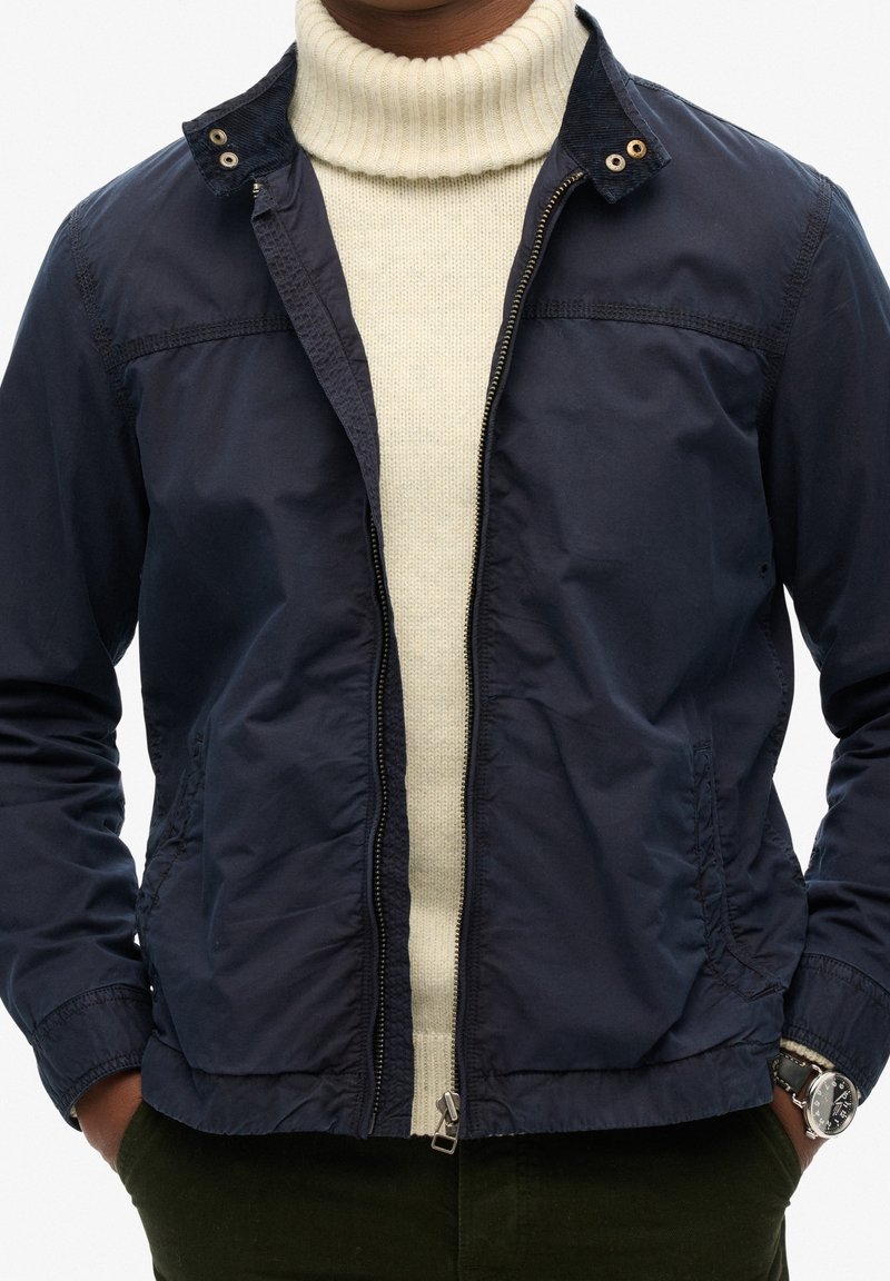 Granatowa lekka kurtka z kołnierzem, frontowym zamkiem błyskawicznym i bocznymi kieszeniami, nałożona na kremowy golf.