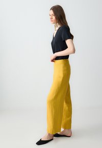 Zwarte korte mouwen V-hals top gecombineerd met hoge taille, wijde broek in felgele linnen. Model draagt zwarte ballerina's.