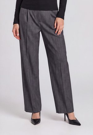 Personne portant un pantalon gris à larges jambes à fines rayures, associé à des escarpins noirs à bout pointu et un haut noir à manches longues.