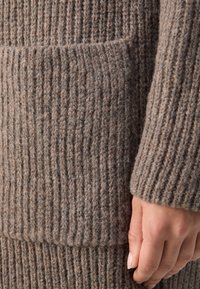 Stickad ulltröja i en taupe-färg med ribbat mönster; har en framficka och en figurnära ärmdesign.