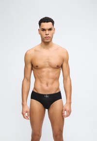 Μυώδης άνδρας στέκεται μπροστά σε άσπρο φόντο, φοράει μαύρα εσώρουχα Calvin Klein, κοιτάζοντας μπροστά με ουδέτερη έκφραση.