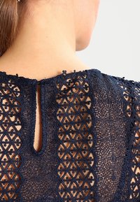 Tummansininen pitsitoppi, jossa on monimutkaisia geometrisia kuvioita, läpikuultavia kohtia ja avainaukollinen kaula-aukko, joka suljetaan nappikiinnityksellä selästä.