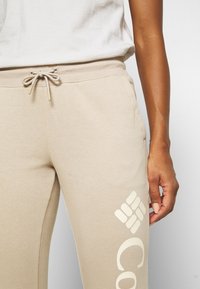 Beige bomullssweatpants med elastisk midjeband och dragsko, med en stor vit logotyp tryckt på vänster lår.