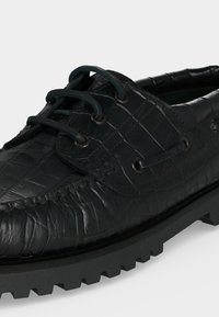 Mocassin en cuir noir avec texture crocodile, design à lacets, détails cousus et semelle épaisse en caoutchouc avec crampons profonds.