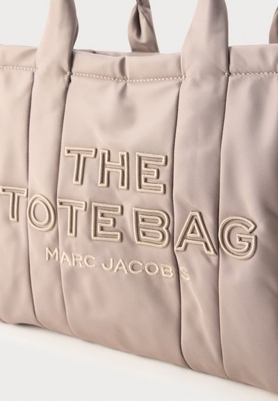 Μπεζ επενδεδυμένη τσάντα tote με κεντητά έντονα γράμματα "THE TOTE BAG" και μικρότερο κείμενο "MARC JACOBS", με δύο υφασμάτινες λαβές.