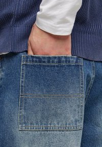 Denim jeans with a faded blue finish, featuring a back pocket and a ribbed navy sweater with a textured knit pattern.