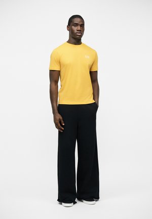 Hombre de pie de frente vestido con una camiseta amarilla de manga corta con logo blanco y pantalones negros anchos con las manos en los bolsillos.