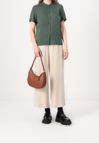 Grön kortärmad skjorta med knappar, beige vida byxor, svarta kraftiga loafers och en brun läderaxelväska. Enkla bakgrund.