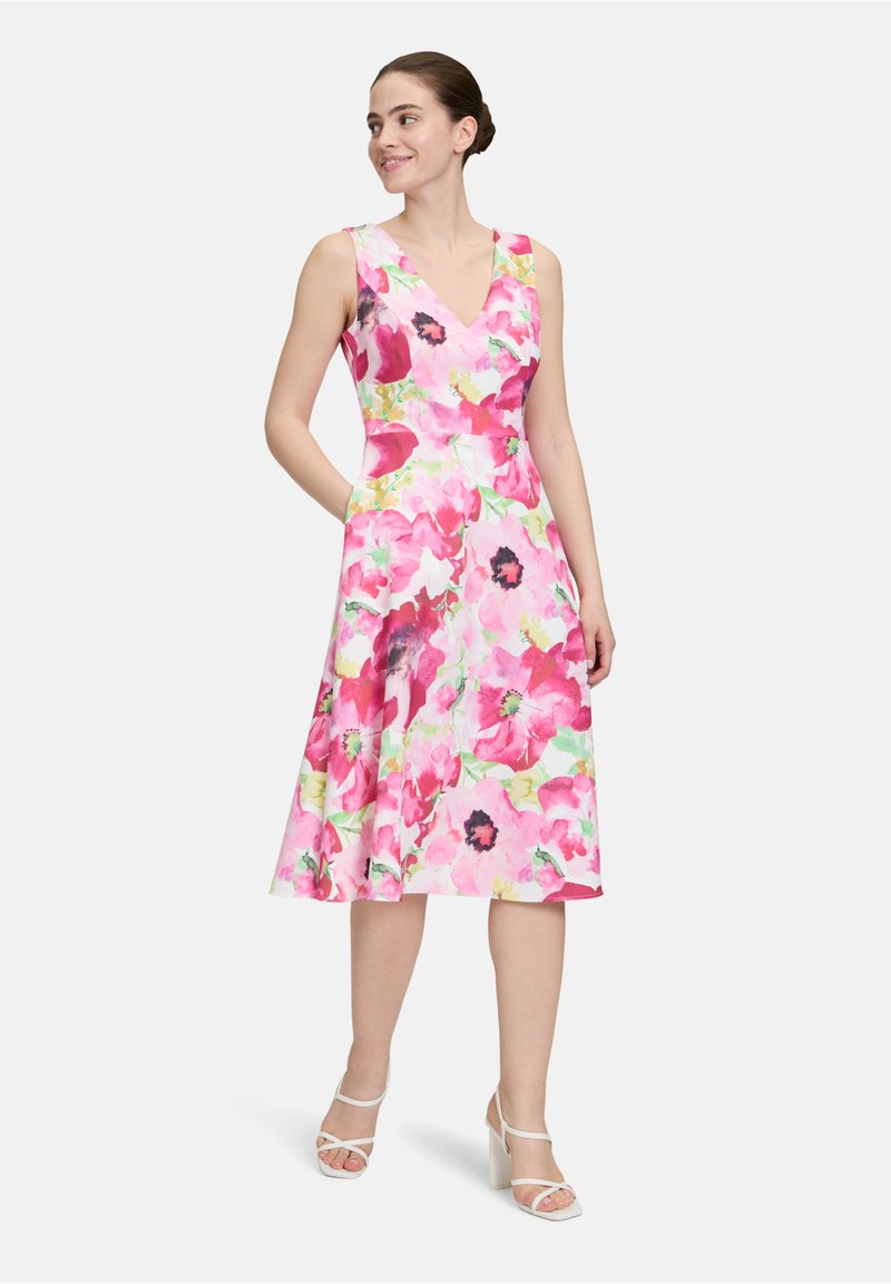 Blumenkleid mit V-Ausschnitt, das ein pinkes und grünes Aquarellmuster aufweist, mit einem weit schwingenden Rock und seitlichen Taschen. Getragen mit weißen Blockabsätzen.