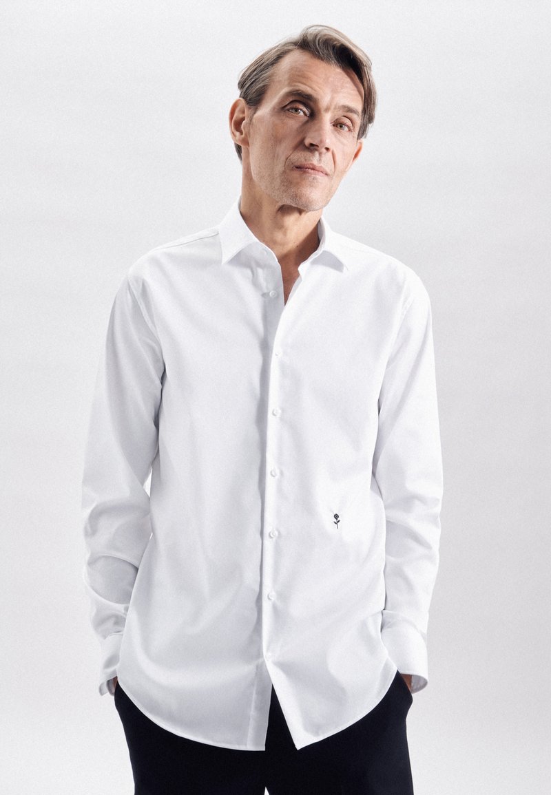 Witte langarmshirt met een spitsere kraag, knopen aan de voorkant en een klein zwart geborduurd rozendetail aan de linkerkant. Glad textiel.