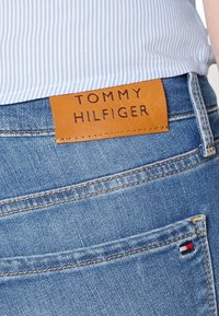 Vaqueros de denim con una etiqueta de cuero marr�ón que dice "TOMMY HILFIGER". Color azul, diseño de pierna recta, costuras visibles y un pequeño acento de bandera.