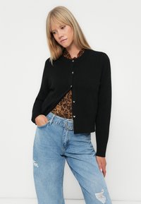 Crna kardigan jakna s okruglim izrezom i gumbima sprijeda, nosi se preko smeđeg gornjeg dijela s leopard uzorkom, uparenih sa svijetlim trapericama sa istrošenim detaljima.