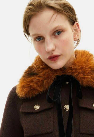 Abrigo de lana marrón con botones dorados, gran cuello de piel sintética naranja y cinta de terciopelo negra atada en el cuello. Tejido texturizado, diseño cuadrado.