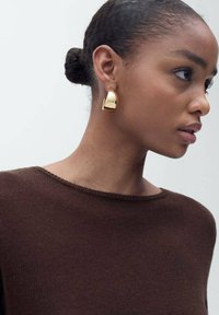 Boucle d'oreille géométrique en or avec une finition polie. Présentée sur une personne portant un pull en tricot marron, mettant en valeur un design lisse et moderne.