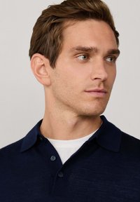 Granatowa koszulka polo wykonana z miękkiego materiału, z klasycznym kołnierzykiem, zapięciem na dwa guziki i lekką teksturą, noszona na białej koszulce w środku.