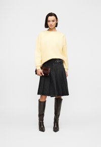 Femme portant un pull crème, une jupe plissée noire avec boucle, tenant une pochette marron foncé, et portant des bottes en cuir marron montantes jusqu'aux genoux.