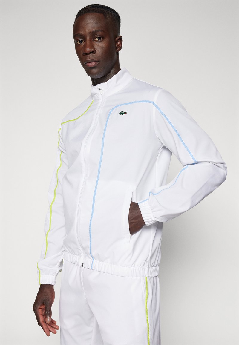Biała kurtka sportowa z niebieskimi i neonowo-żółtymi akcentami, z zamkiem z przodu, elastycznymi mankietami i kieszeniami, wykonana z lekkiego materiału.