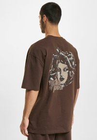 Brun oversized t-shirt lavet af bomuld med et grafisk print af en kvinde med slanger på ryggen, med teksten "THE RIDER OF MURDER."
