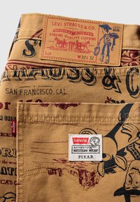 Denim de Levi's con base beige, que presenta gráficos impresos en rojo y negro y una etiqueta de cuero marrón. Incluye una etiqueta blanca con el logotipo de "Pixar".