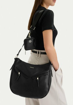 Kobieta w czarnej koszulce z krótkim rękawem i beżowych spodniach niesie dużą czarną torbę na ramię marki Lasocki z małą przymocowaną czarną saszetką.