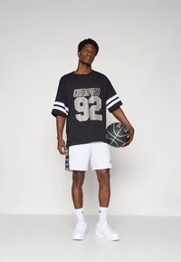 T-shirt preta oversized com estampa cinza "CHAMPION 92" e riscas brancas. Calções brancos com logótipos laterais e bola de basquetebol preta. Ténis brancos.