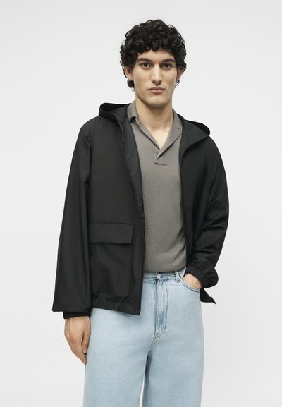 Νεαρός άνδρας με σγουρά μαλλιά, φοράει μαύρο μπουφάν με κουκούλα πάνω από γκρι πόλο και ανοιχτό μπλε τζην, στέκεται με το ένα χέρι στην τσέπη.